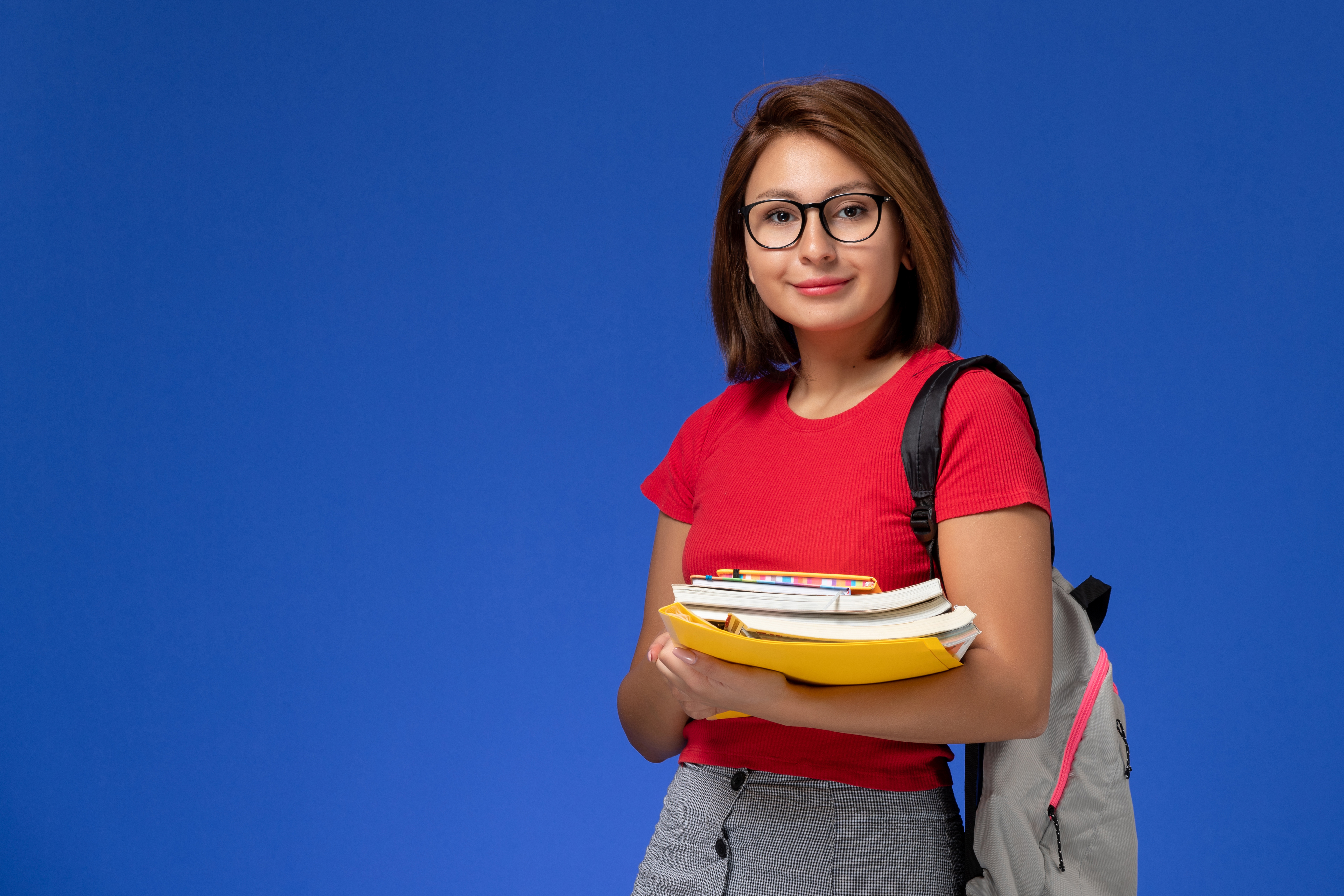front view female student red shirt with backpack holding books files smiling blue wall